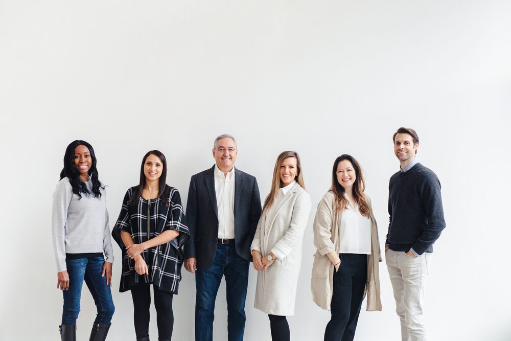 Team of six standing in a row against a white wall in a bright studio