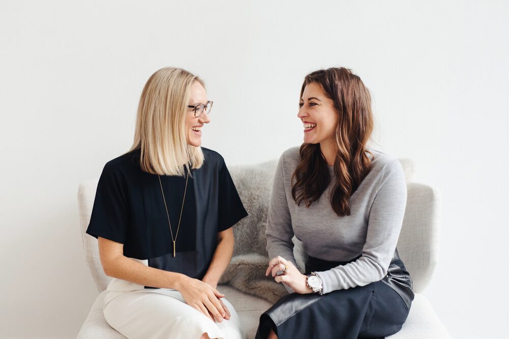 Two women laughing together on a white sofa