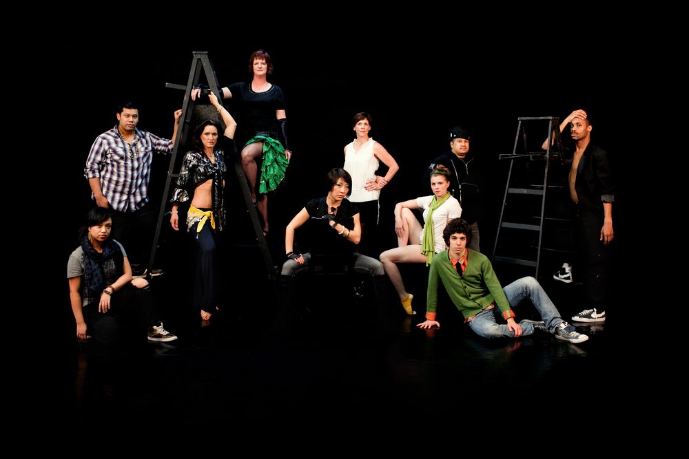 Cast of performers posed on a dark stage with ladders