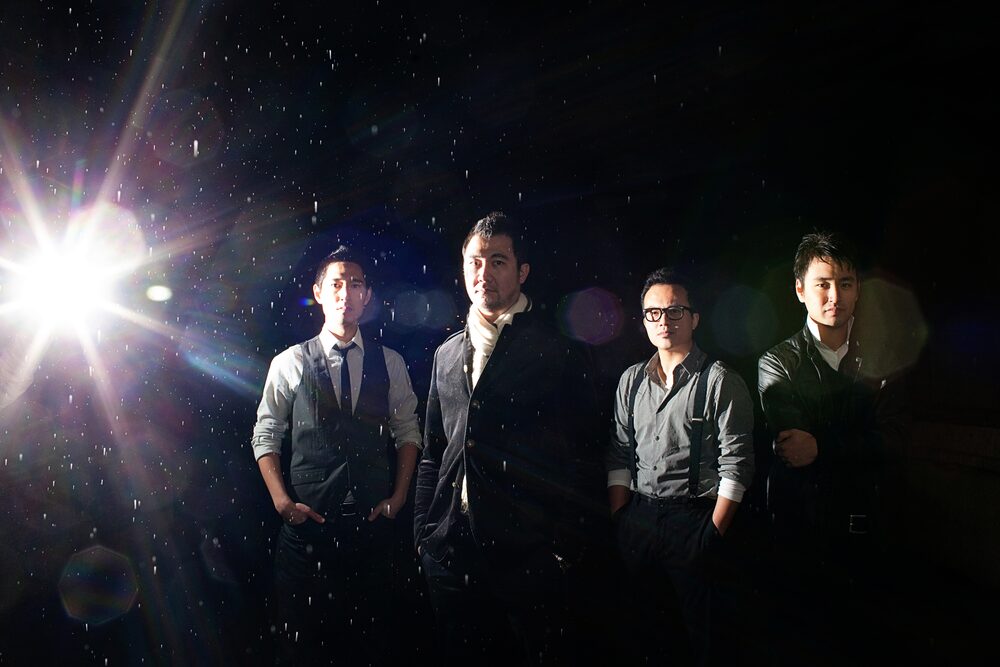 Four-piece band standing in the dark with a lens flare behind them