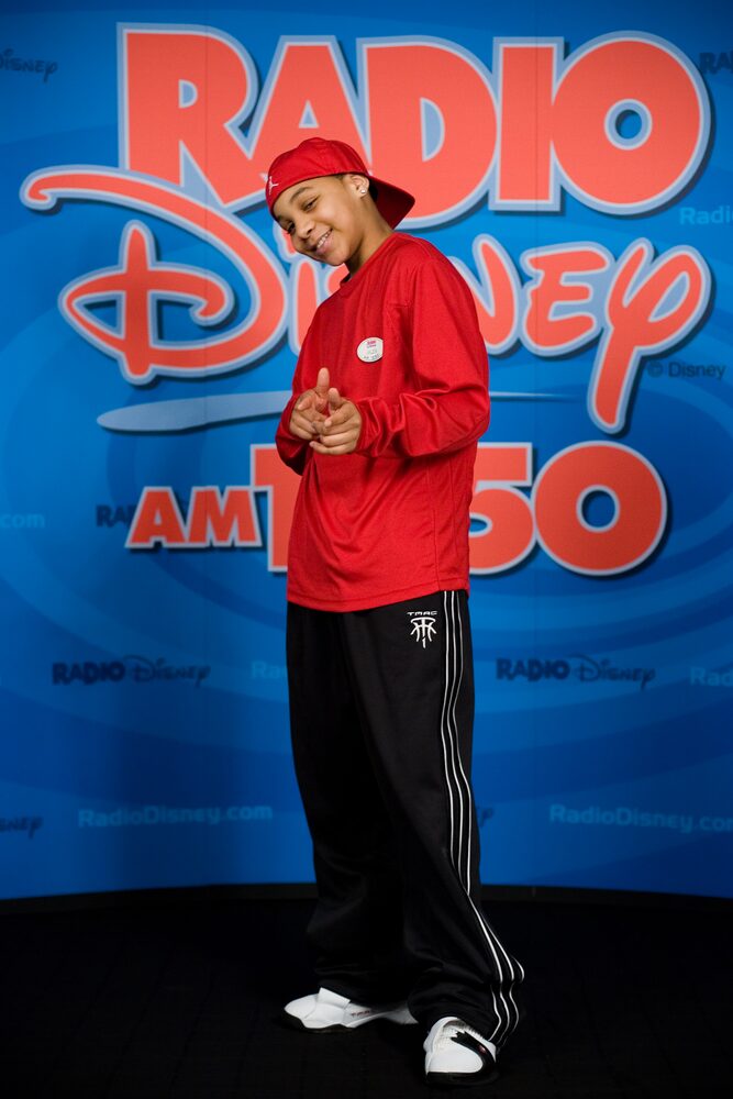 Young performer in a red hoodie and cap in front of a radio station backdrop