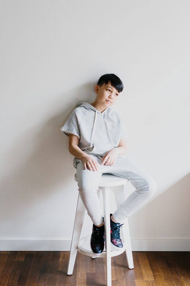 Boy in a grey hoodie seated on a white stool against a white wall