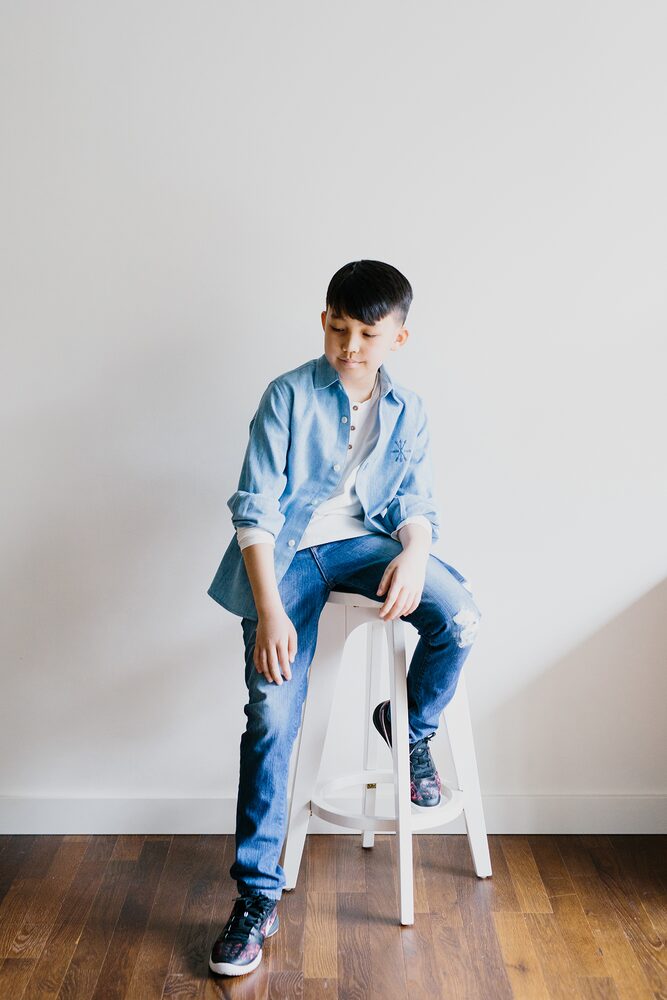 Boy in a denim shirt seated on a white stool against a white wall