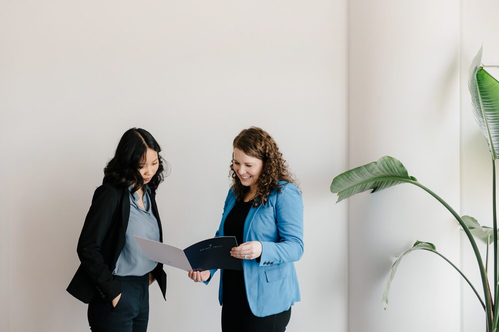 Two women in blazers reviewing a document beside a houseplant