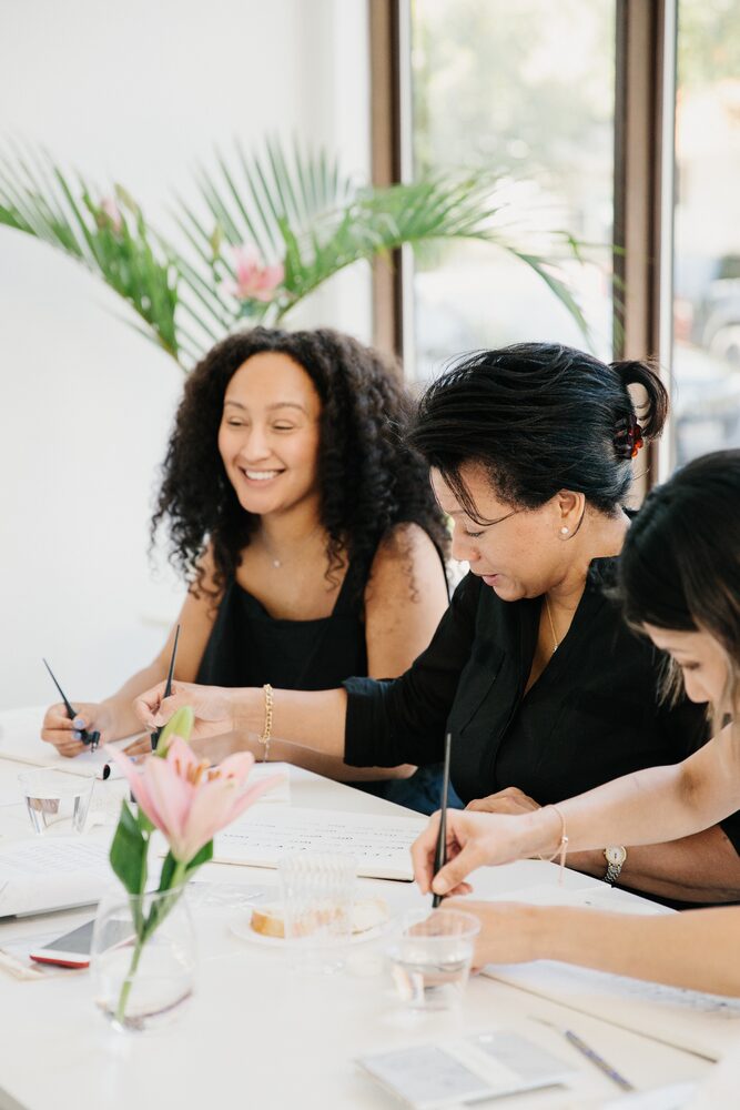 Two women writing at a workshop table with a single flower in a vase