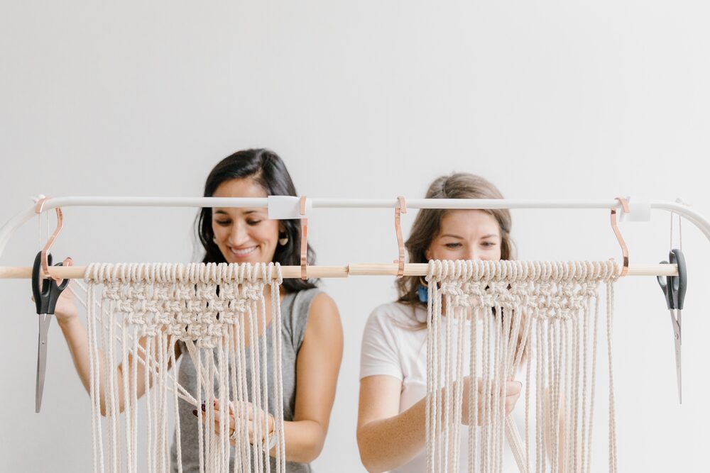 Two women weaving a macramé wall hanging on a dowel