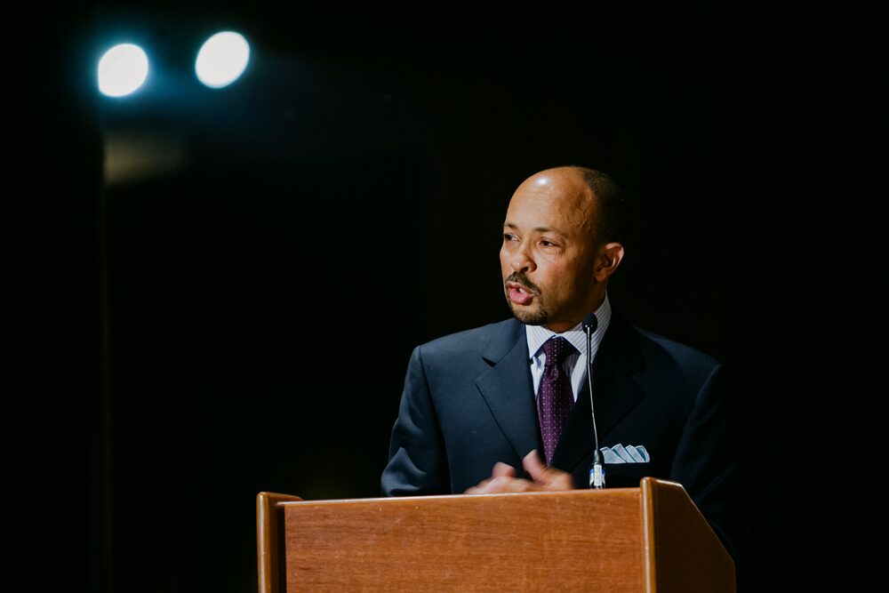 Speaker at a podium lit by stage lights against a dark background