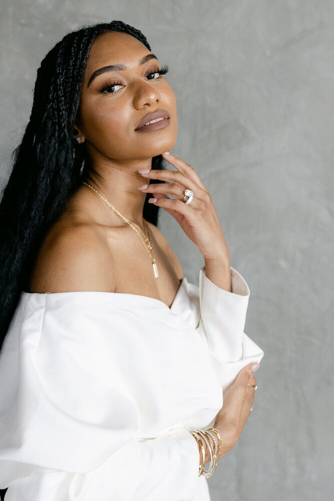 Woman with long braids in an off-the-shoulder white top, hand at her chin