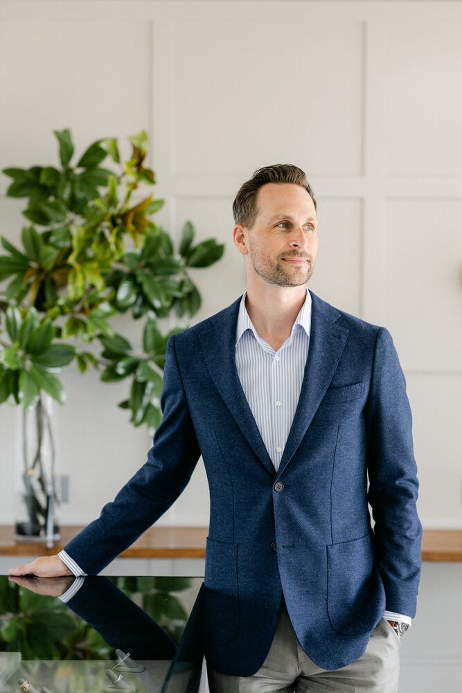 Man in a navy blazer standing beside a fiddle-leaf fig in a bright room