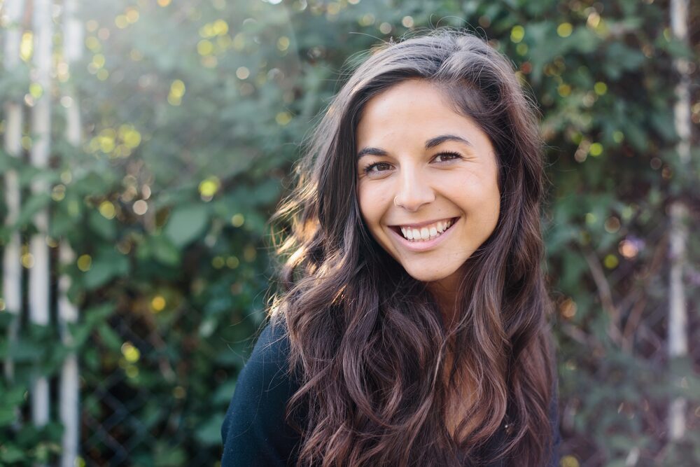Woman with long dark curls smiling in front of green foliage