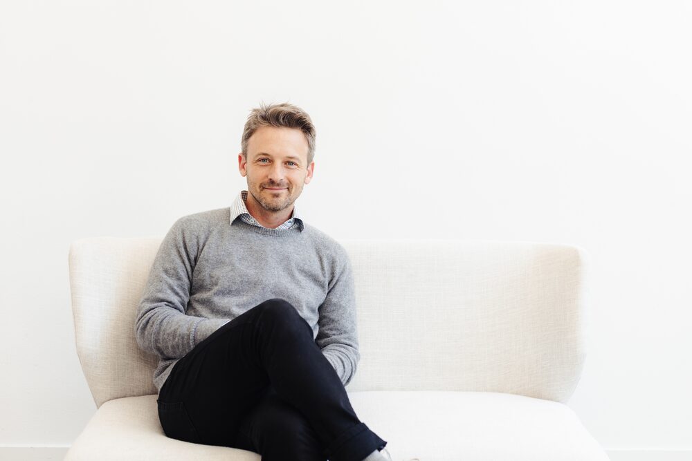 Man in a grey sweater seated on a white sofa in a bright room
