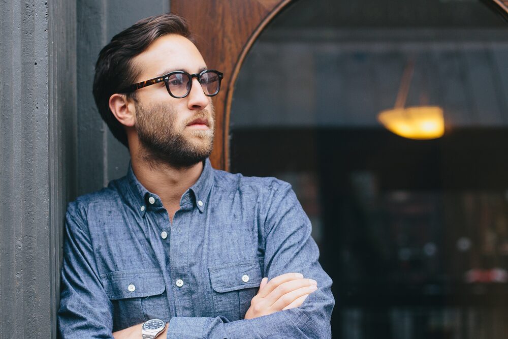 Man with glasses and a beard in a chambray shirt, arms crossed, by a window