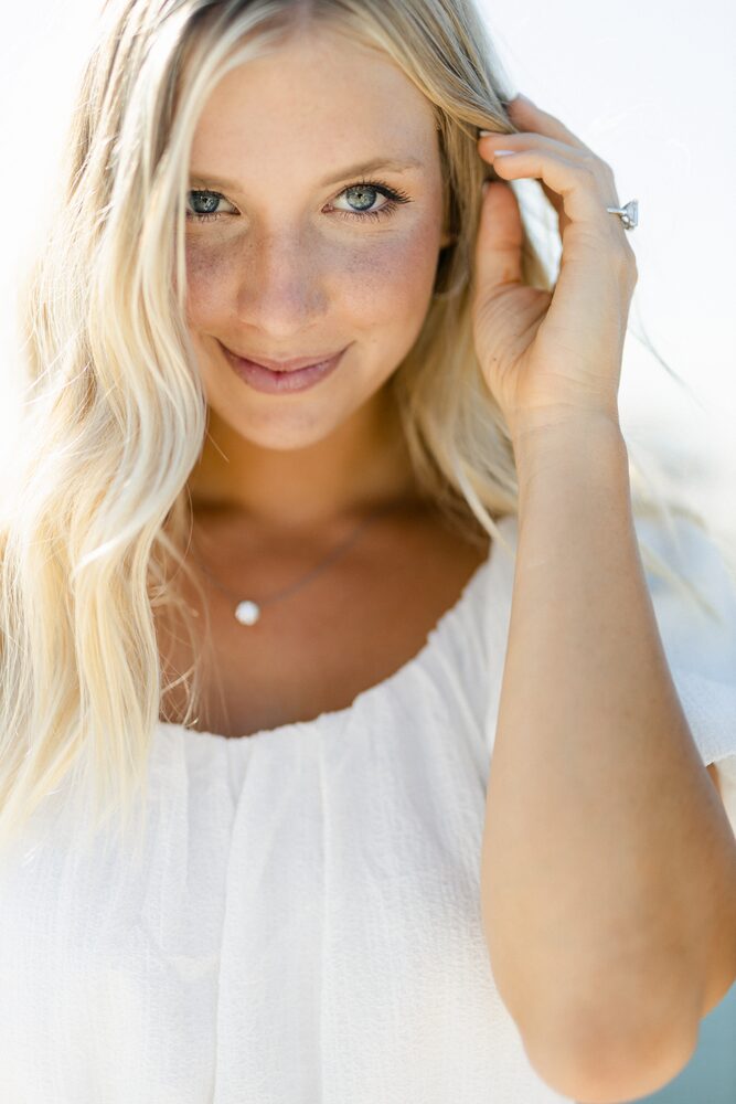 Woman with long blonde hair in a white top, brushing hair from her face in soft light