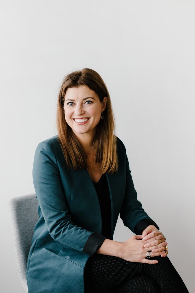Woman with dark hair in a teal blazer seated with hands clasped, smiling
