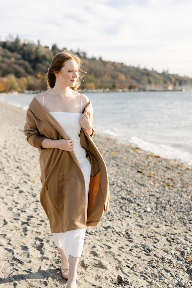 Woman in a long camel coat walking along a pebbled beach