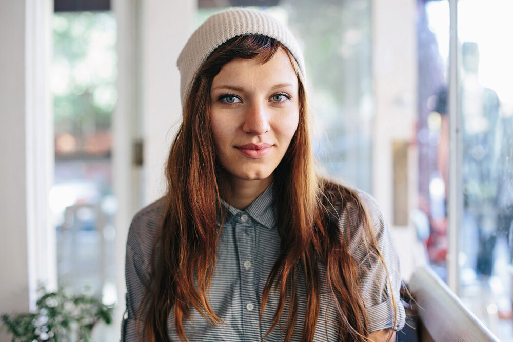 Woman in a knit beanie and denim shirt seated by a sunlit window