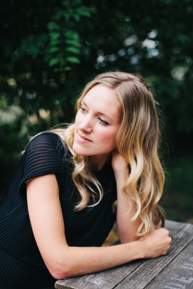 Woman with long blonde hair in a black top resting her arms on a wooden table among leaves