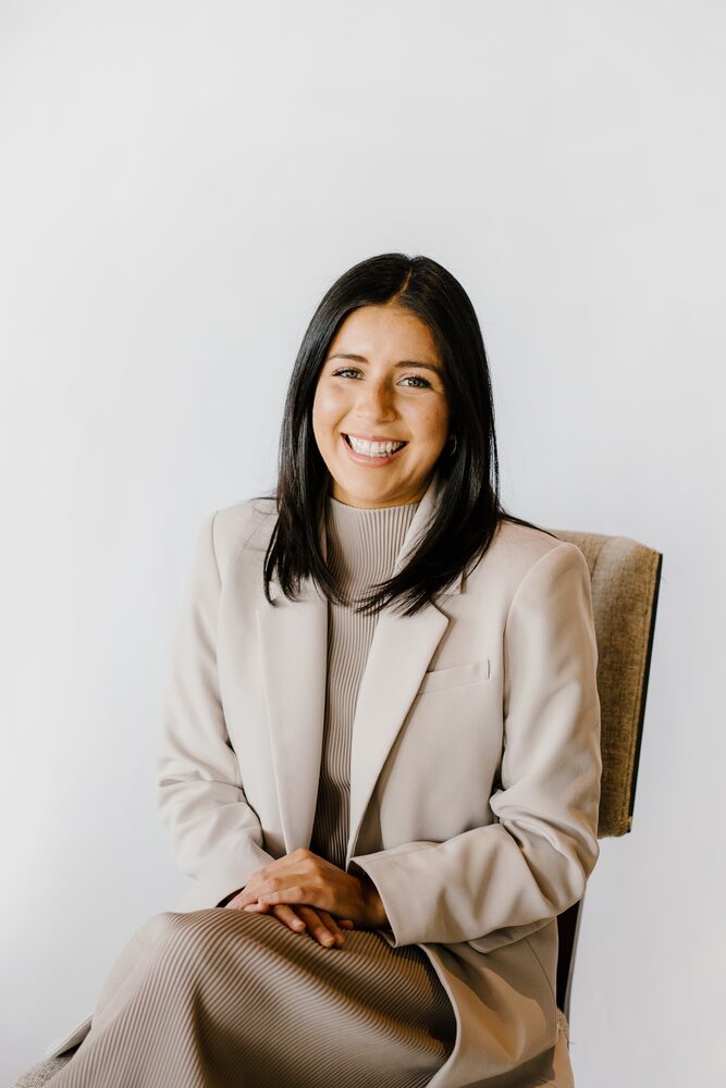 Woman with dark hair in a cream blazer seated and smiling