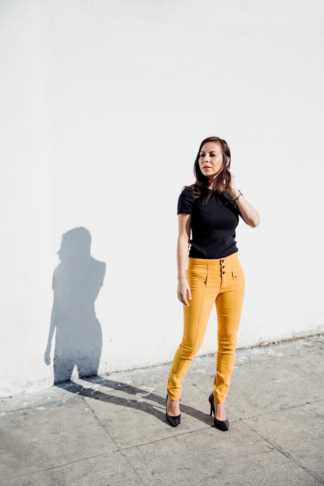 Woman in a black top and mustard trousers standing against a sunlit white wall