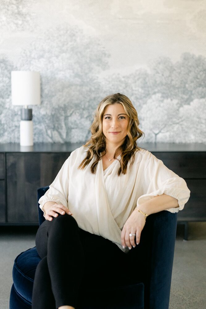 Woman in a white blouse seated in a navy armchair in front of a wallpaper mural