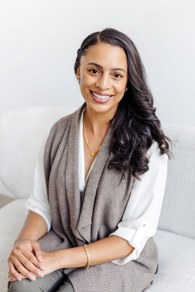 Woman in a white blouse and grey wrap seated with hands clasped, smiling