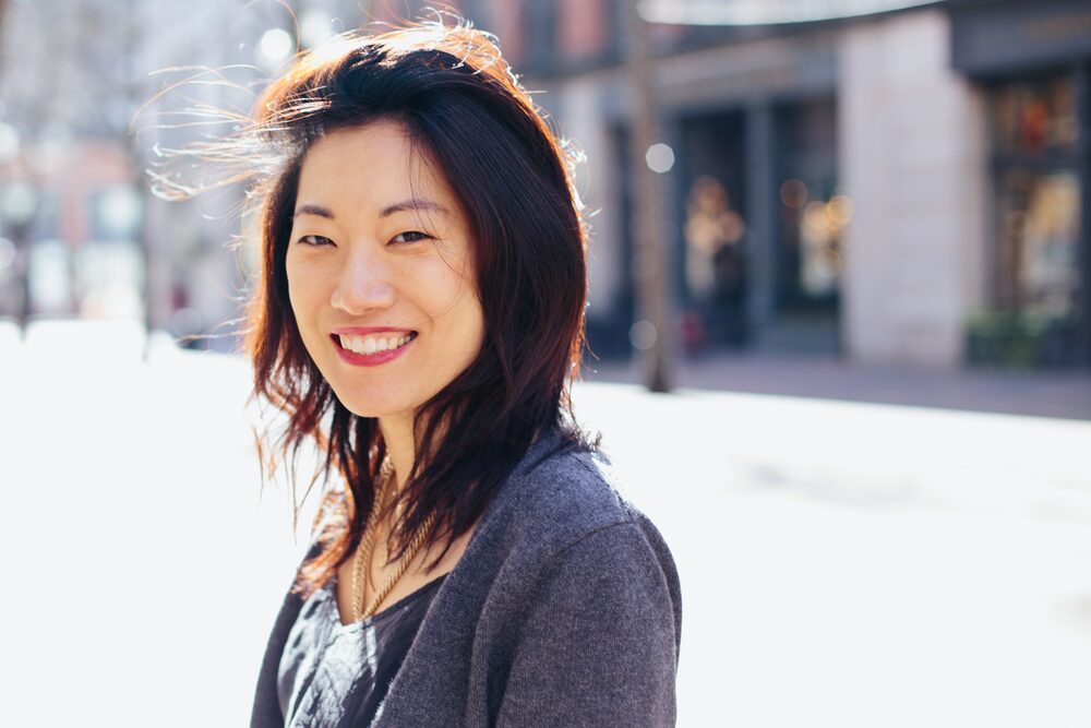 Woman with short dark hair in a grey cardigan smiling on a city sidewalk