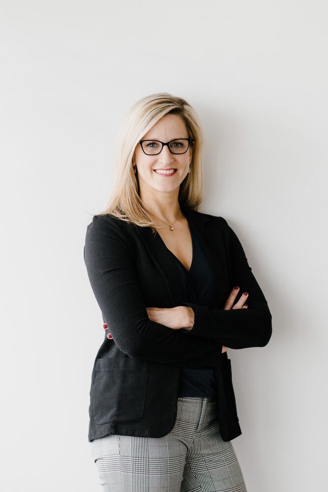 Woman with glasses in a black blazer, arms crossed, against a white wall