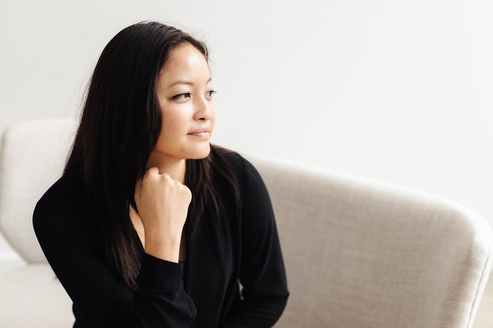 Woman with long dark hair in a black top seated on a light sofa, looking to the side
